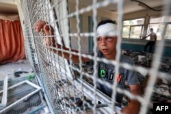 Seorang anak laki-laki yang terluka berdiri di dekat kisi-kisi jendela di ruang kelas yang hancur tempat orang-orang berlindung yang sebelumnya terkena pengeboman Israel, di Kamp Nuseirat di Jalur Gaza tengah. (Foto: AFP)