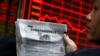 FILE - A man reads a newspaper as he looks at an electronic board displaying stock prices at a brokerage house in Beijing, Dec. 5, 2016.