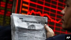 FILE - A man reads a newspaper as he looks at an electronic board displaying stock prices at a brokerage house in Beijing, Dec. 5, 2016.