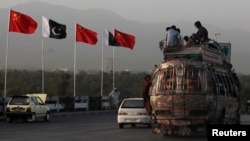 People sit on top of a bus as they go past flags of Pakistan and China that are displayed along a road.