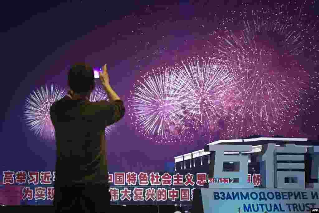 2018年6月9日,在山东青岛举行的上海合作组织(SCO)峰会期间,礼花绽放,焰火绚丽,一名男子用手机拍摄照片。此次上合组织青岛峰会在官媒渲染下盛况空前,中国为与会国代表举行了欢迎宴会,还有文艺演出和焰火表演。有一些推特用户以此和同一时段举行的工业7国峰会做比较,赞扬G7峰会之简朴,批评青岛峰会之奢华。有人说:“西方富国开穷会,东方穷国开富会。”