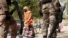 In this photo taken Wednesday, April 8, 2015, a woman walks past Nigerian Soldiers at a checkpoint in Gwoza, Nigeria, a town newly liberated from Boko Haram. Each day brings new reports of atrocities, with mass graves being discovered in towns seized back from the militants who had set up a so-called “Islamic caliphate” across a great swath of northeast Nigeria. Boko Haram's nearly 6-year-old Islamic uprising in northeast Nigeria that has killed thousands — a reported 10,000 just last year — and forced more than 1.5 million from their homes. (AP Photo/Lekan Oyekanmi)