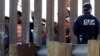 ARSIP – Sekumpulan kaum migran dari Amerika Tengah memandang melalui sela-sela dinding perbatasan saat petugas U.S. Customs and Border Protection, kanan, bersiaga di sisi yang berlawanan, Minggu, 25 November 2018 (foto: AP Photo/Gregory Bull)