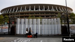 Dua pejalan kaki mengenakan masker di tengah pandemi virus corona (COVID-19), melintas di depan Stadion Nasional di Tokyo, tempat penyelenggaraan Olimpiade Tokyo 2020 dan Paralimpiade, Tokyo, Jepang, 7 Juli 2021. 