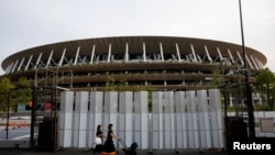 အိုလံပစ်ပွဲကျင်းပမည့် တိုကျိူမြို့ National Stadium အရှေ့မှ မျတ်နှာ၊ နှာခေါင်းစည်း အကာအကွယ်တွေနဲ့ ဖြတ်လျှောက်သွားသူများ (ဓာတ်ပုံ - Reuters)