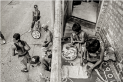 យុវជនខ្មែរមួយក្រុមជុំគ្នានៅក្រោយអគារមួយ ខណៈស្ត្រីម្នាក់និងកុមារម្នាក់កំពុងអង្គុយកាត់ក្រណាត់ក្រមា នៅអគារគេហដ្ឋានក្នុងតំបន់ជាយក្រុង Chicago រដ្ឋ Illinois កាលពីដើមទសវត្សរ៍ ១៩៩០។