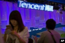 FILE - A woman uses her smartphone near a booth for the Chinese Internet company Tencent at the Global Mobile Internet Conference in Beijing, China, April 29, 2015.