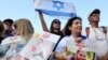 Orang-orang membawa poster yang memuat gambar sandera Israel yang baru dibebaskan, Yarden Bibas, serta kedua anaknya, Kfir dan Ariel, saat mereka menonton pembebasan sandera Israel di Jalur Gaza di layar di lapangan sandera di Tel Aviv pada 1 Februari 2025. (Foto: AFP)