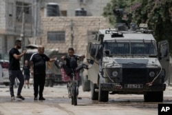 Seorang remaja mengendarai sepeda melintas di samping kendaraan militer Israel yang sedang melancarkan operasi militer di Jenin, Tepi Barat, Rabu, 28 Agustus 2024. (Foto: Majdi Mohammed/AP Photo)