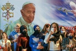 Para perempuan menunggu di luar Katedral Khaldea Saint Joseph, di Baghdad, Irak, Sabtu, 6 Maret 2021, di mana Paus Fransiskus, yang digambarkan pada poster raksasa di punggung mereka, sedang mengadakan misa. (Foto: AP)